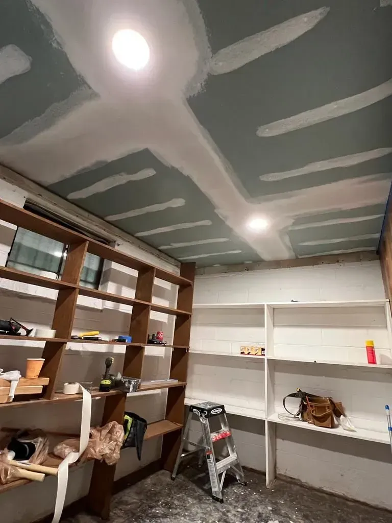 Unfinished storage room with shelves, white workbenches, ladder, and a ceiling under renovation with taped seams.