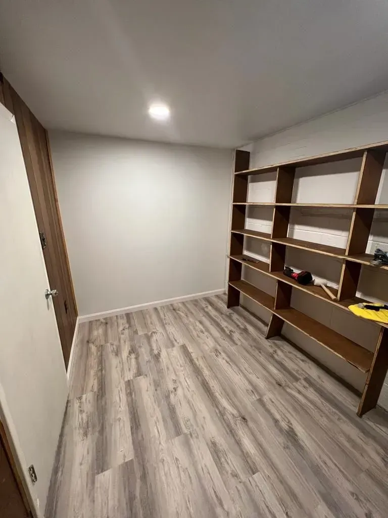 Empty white room with gray wood flooring and built-in wooden shelves on the right.