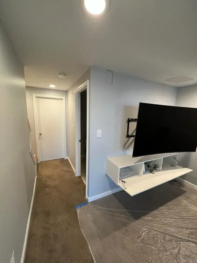Modern hallway leading to a white door, with a wall-mounted TV and floating shelf in the foreground.