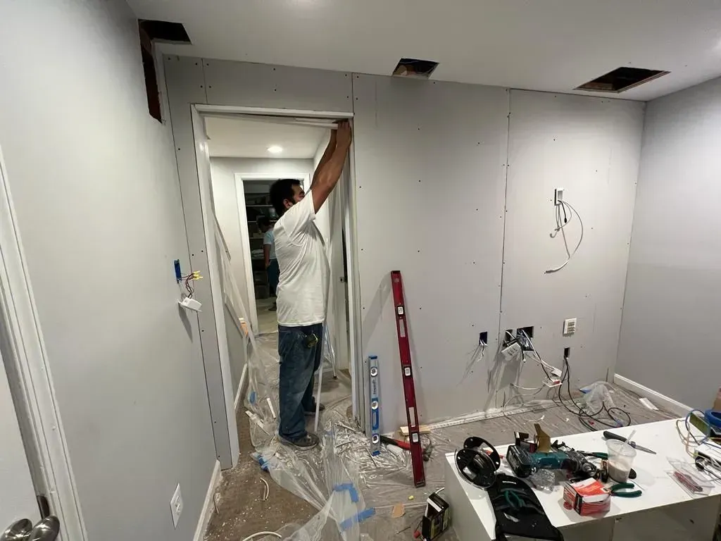 Worker installing drywall in a room under renovation, with tools and plastic sheeting on the floor.