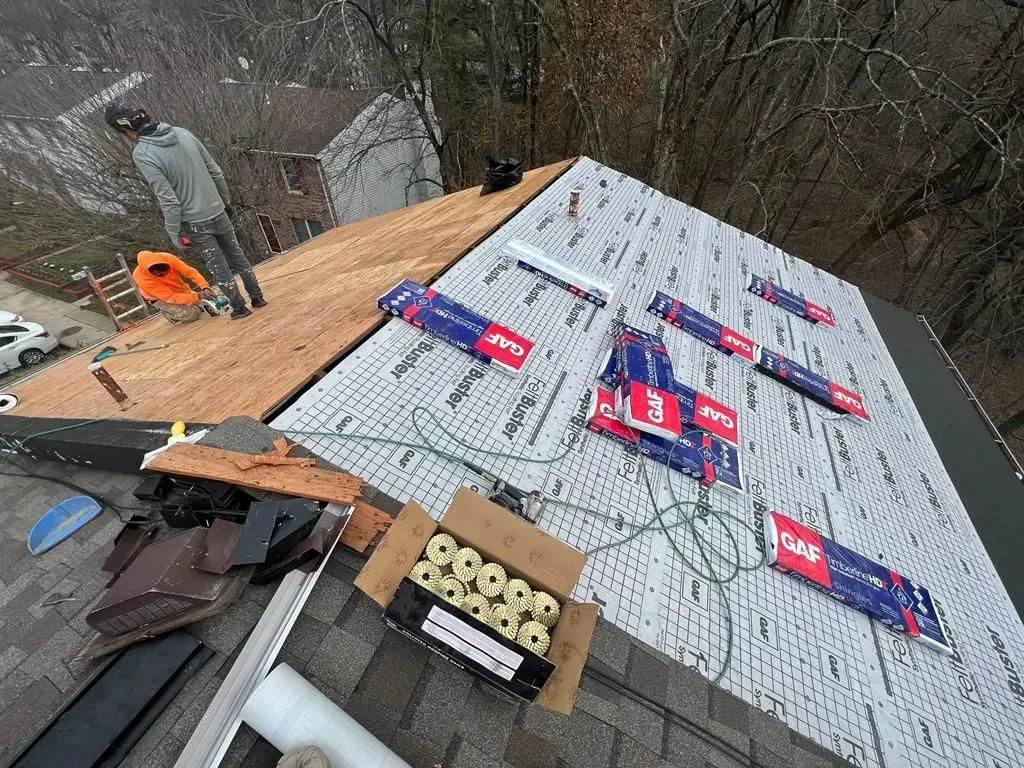 Roofer installing white underlayment and shingles on a steep roof with tools and materials nearby
