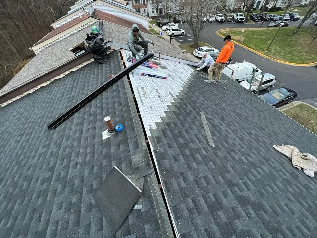 Workers repairing a gray shingled roof with tools and materials near parked cars below.