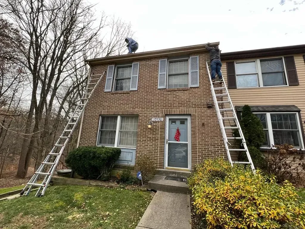 Two ladders lean against a brick house with holiday decorations on the front door.