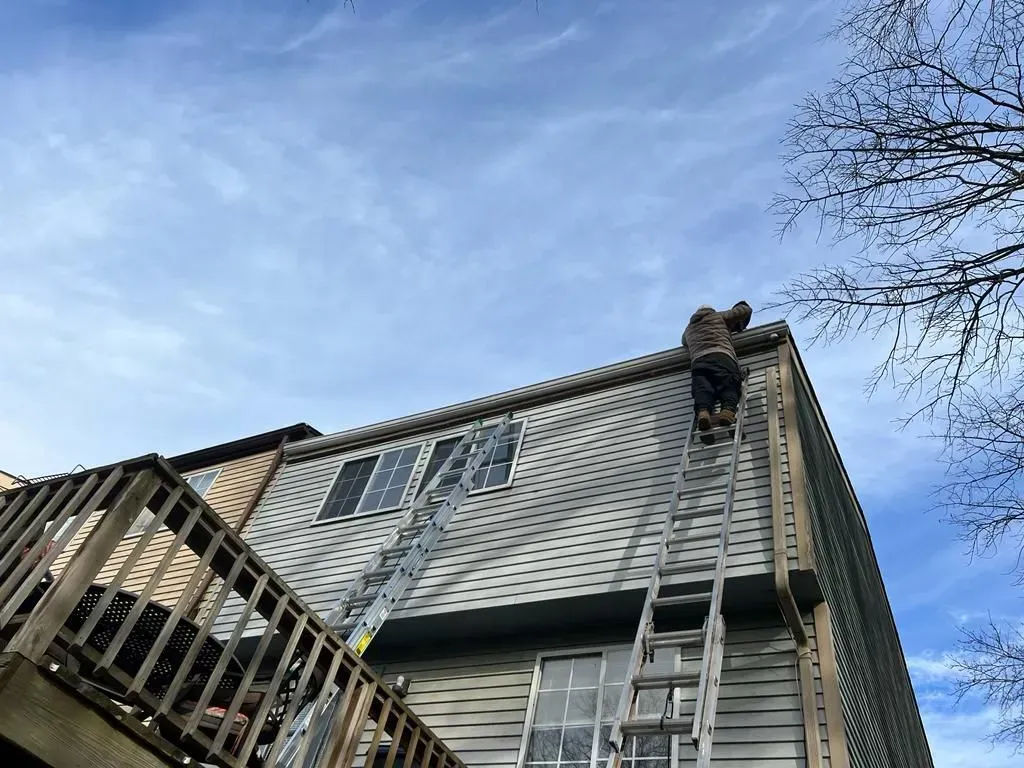 Damaged house roof with missing shingles and a ladder against the siding under a blue sky