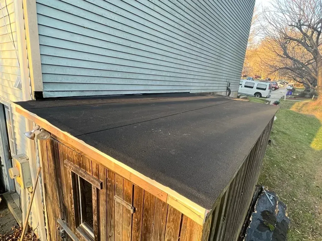 Black roof on a wooden structure beside a house, with autumn trees and a street in the background
