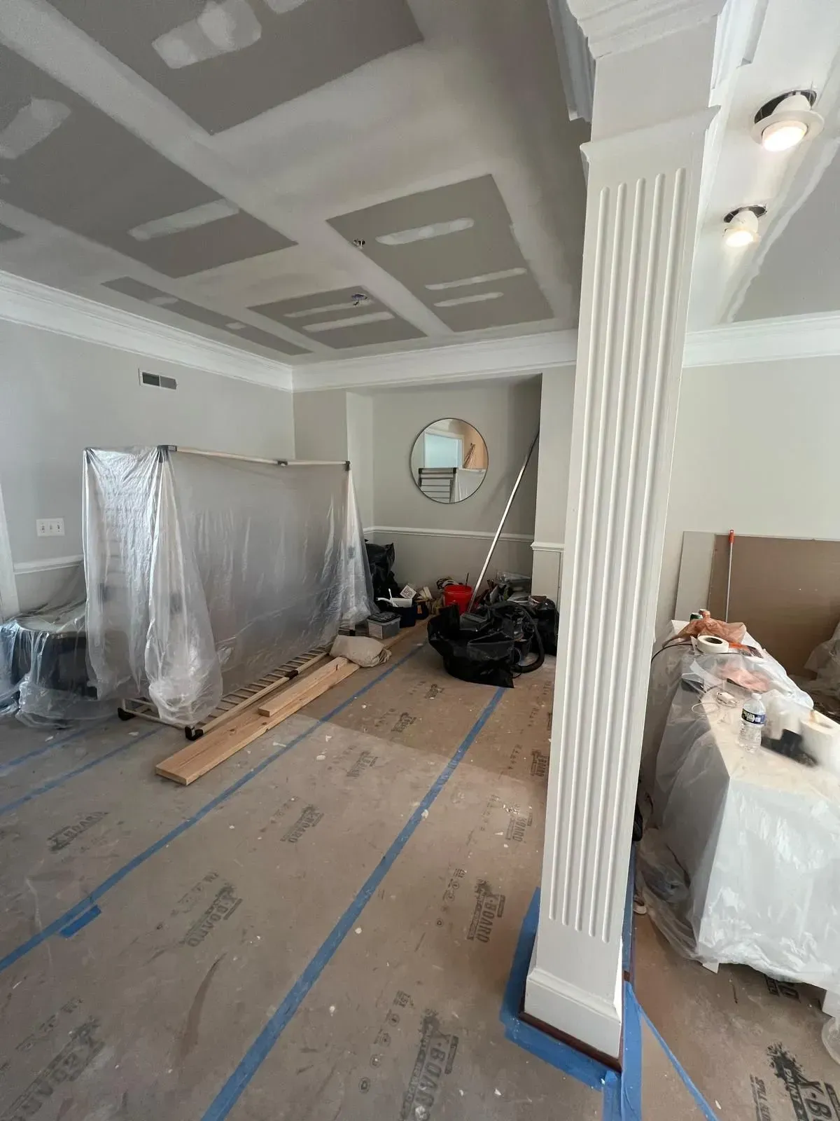 Renovation in progress in a living room with covered furniture, tools, and taped floor paths