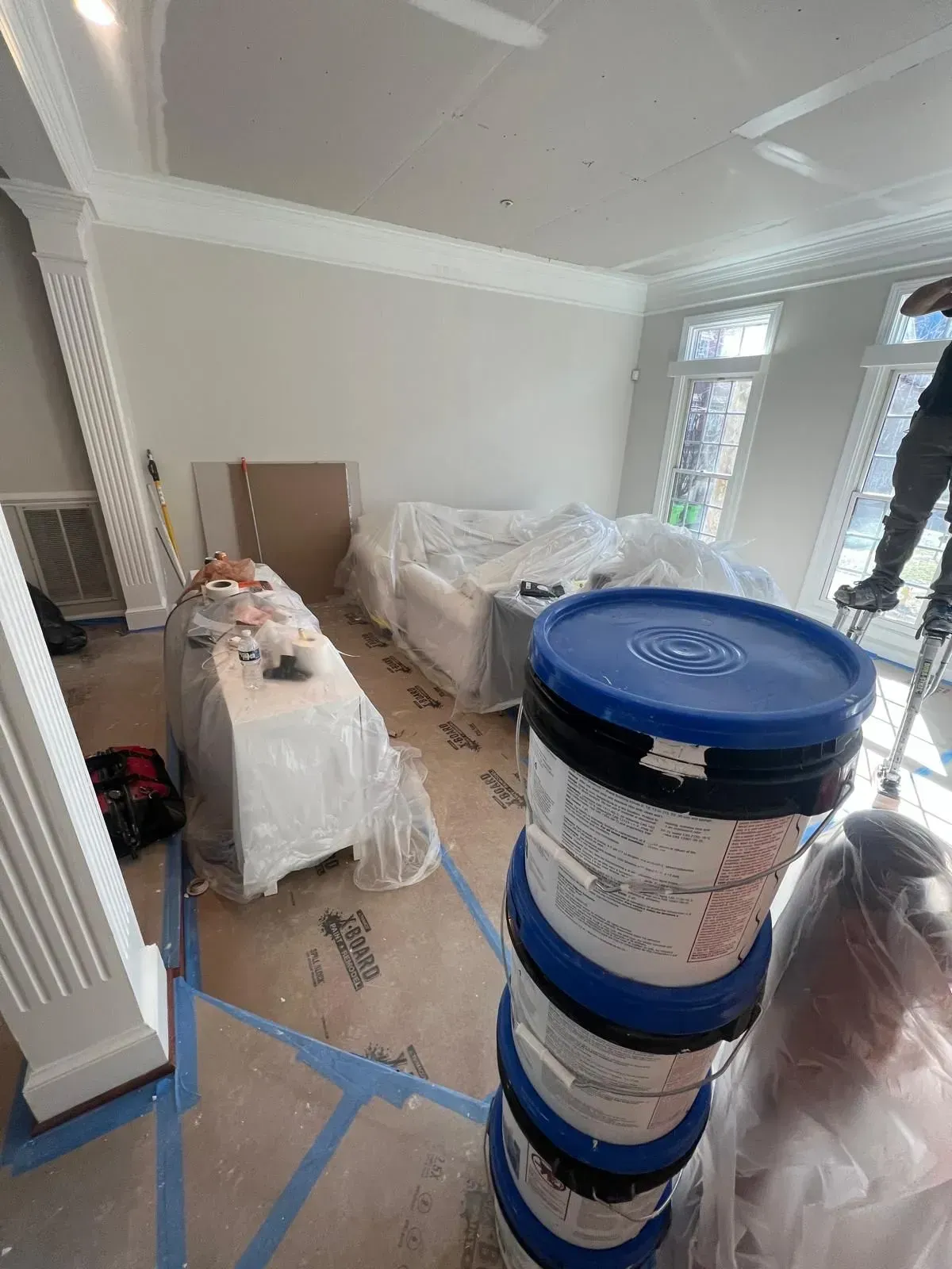 Renovation room with stacked paint buckets, plastic-covered furniture, and blue tape on the floor