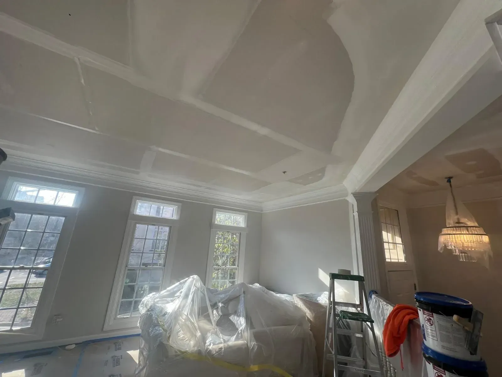 Living room under renovation with plastic-covered furniture, ladder, and chandelier under a coffered ceiling