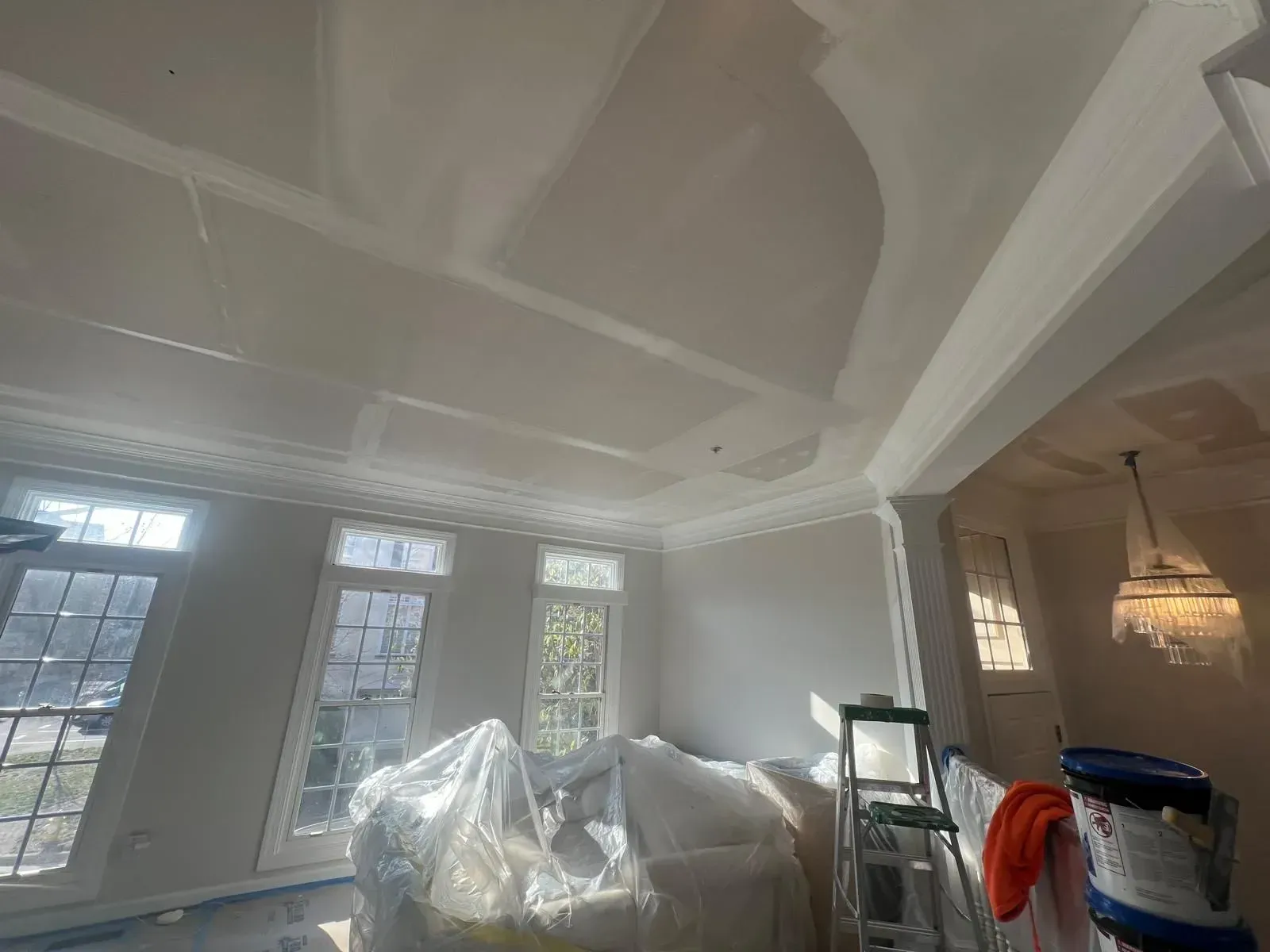 Living room under renovation, with plastic-covered furniture, ornate ceiling moldings, and large windows