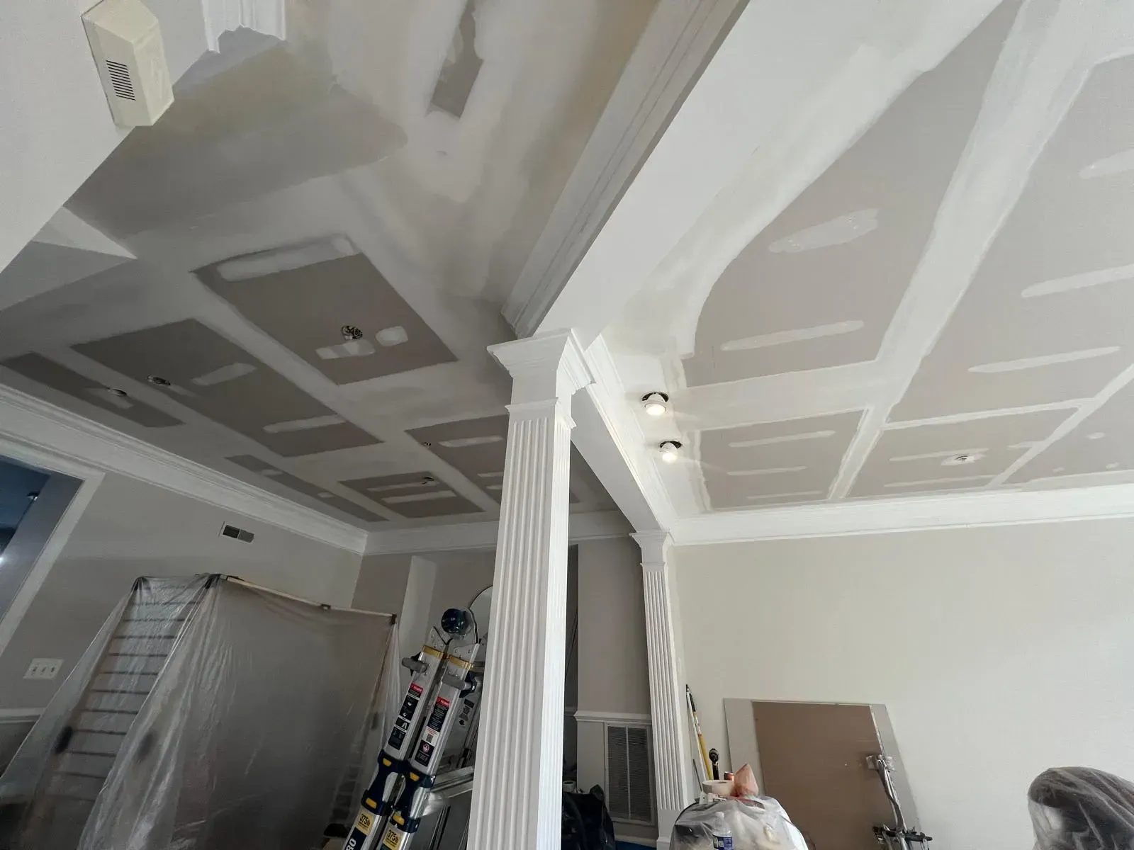 Renovation site with taped drywall ceiling, columns, ladder, and plastic-covered furniture in a room