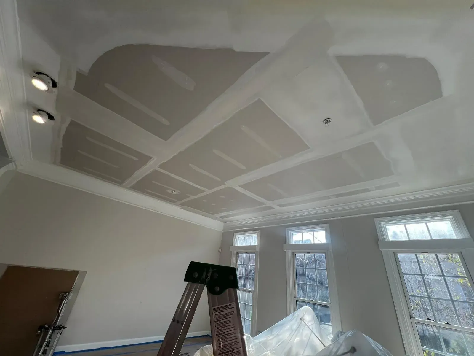 Partially finished ceiling with drywall seams, ladder below, and windows letting in daylight