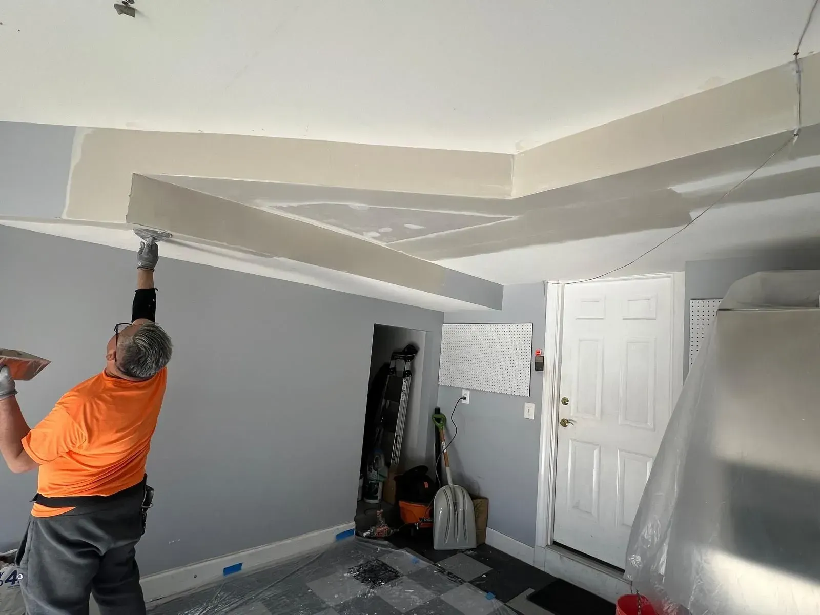 Person in orange shirt painting a gray room, reaching up to cut in the ceiling edge.