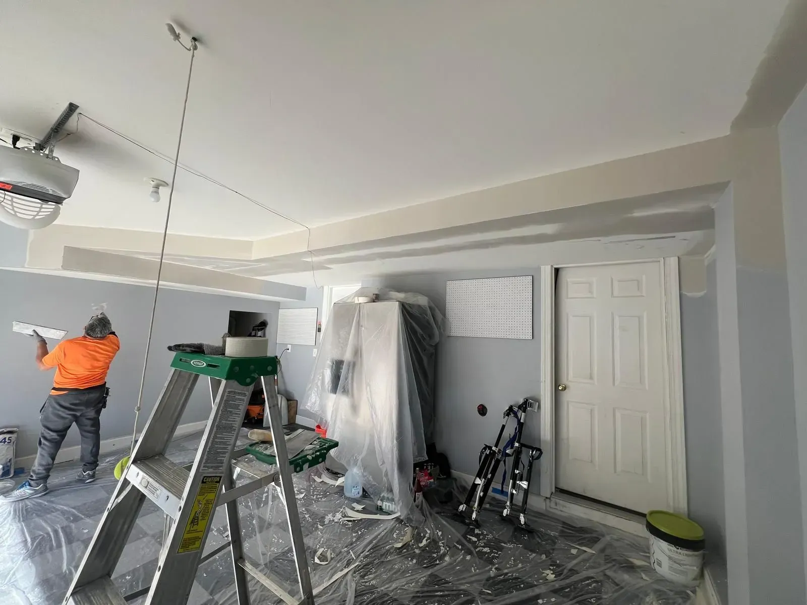 Garage under renovation with worker on ladder, tools, covered furniture, and open wall panels