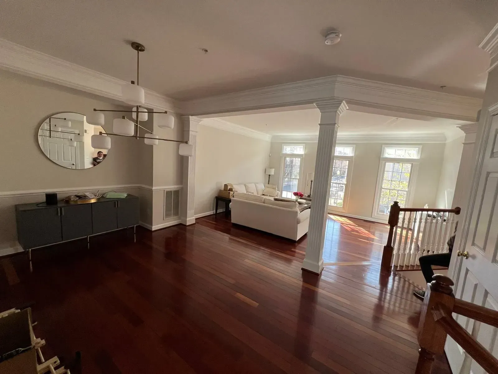 Spacious living room with polished wood floors, white columns, a round mirror, and bright windows overlooking a deck