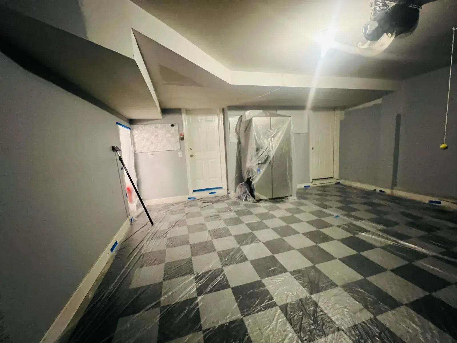Empty basement room with gray walls, checkered floor, exposed ceiling, and hanging work light.