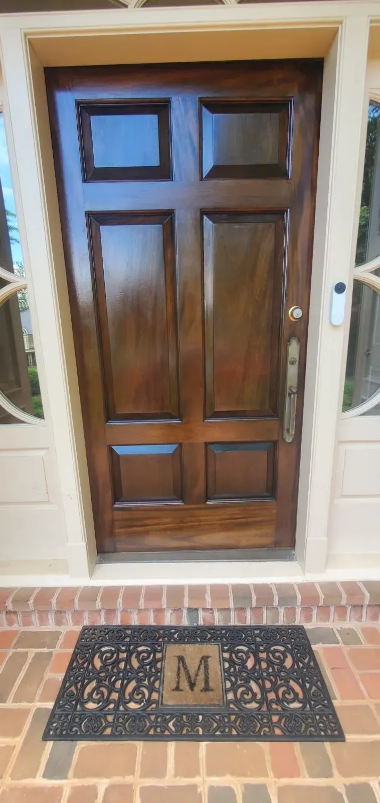 Wooden front door with sidelights and a monogrammed welcome mat on a brick porch.