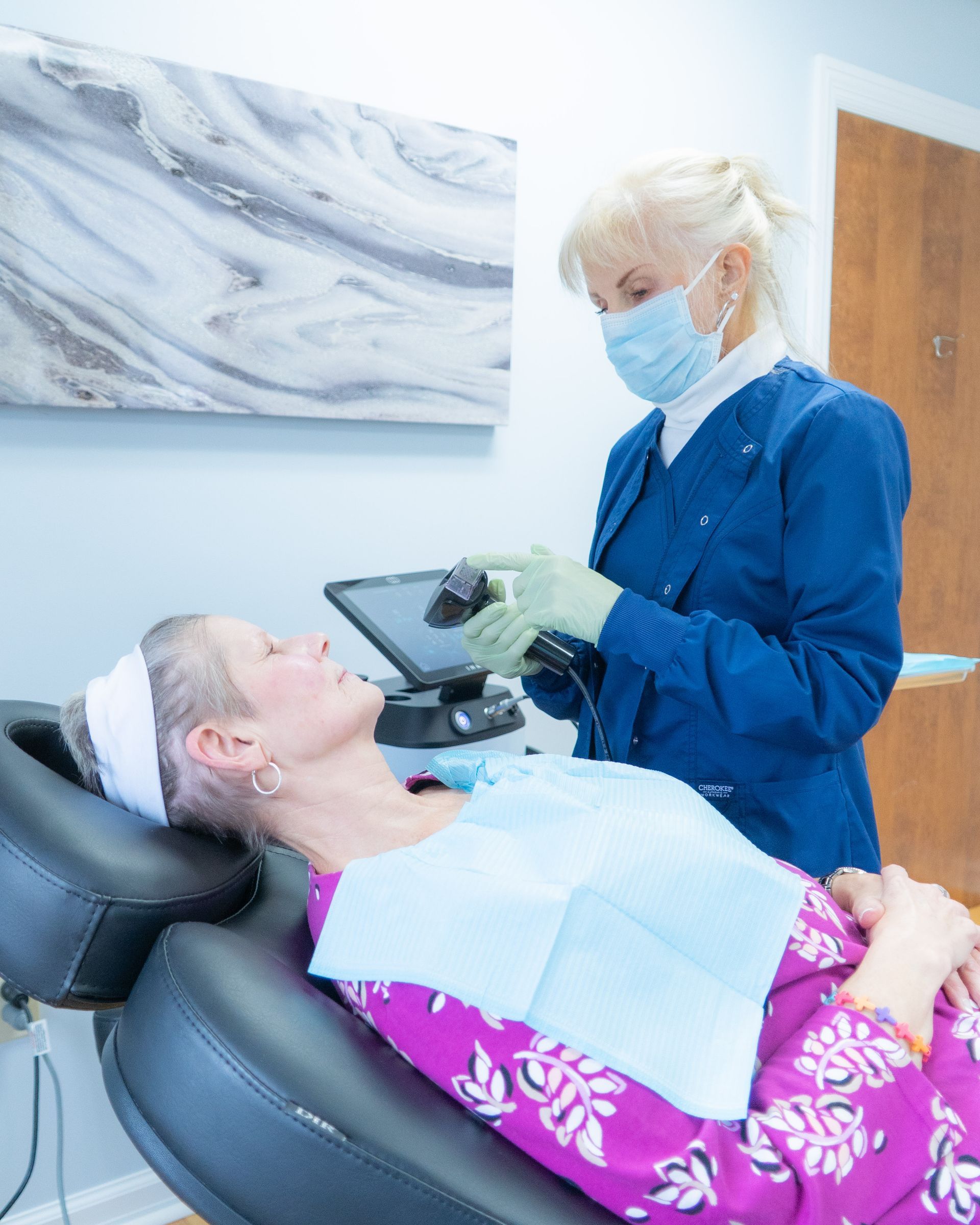 Dentist in blue scrubs examining a patient in a dental chair; medical setting.