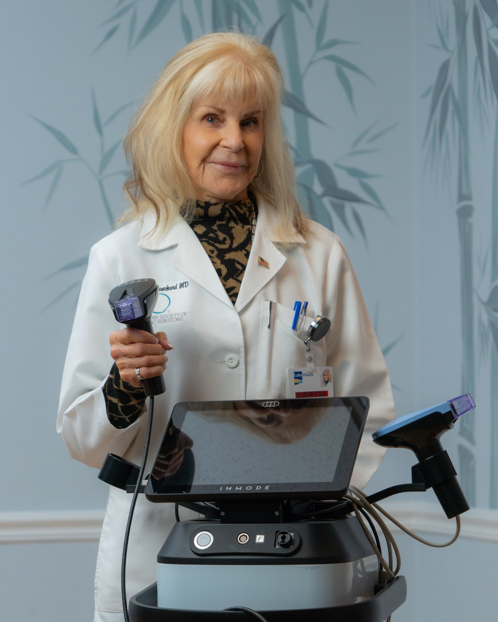 Woman in lab coat holds device, stands near machine. Light blue bamboo wallpaper.