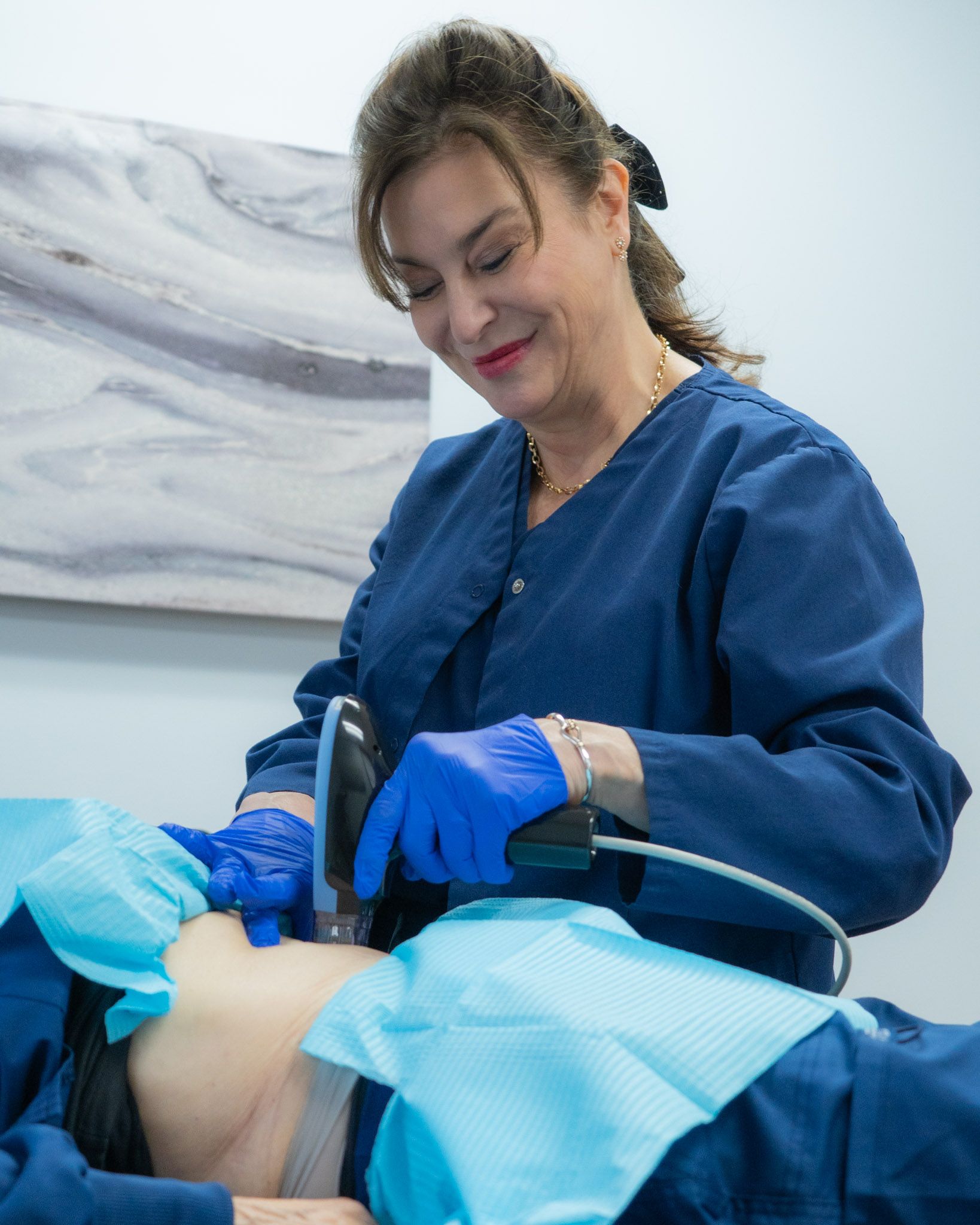 Woman in scrubs using a medical device on a patient's abdomen in a clinic setting.