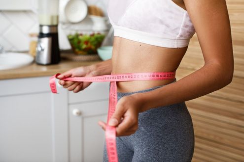 Woman in athletic wear measuring her waist with a pink tape measure in a kitchen setting.