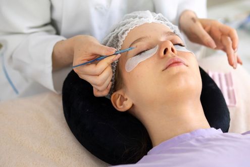 Woman receiving eyelash extensions. Technician using tools, eye patches, and cap, in a salon setting.