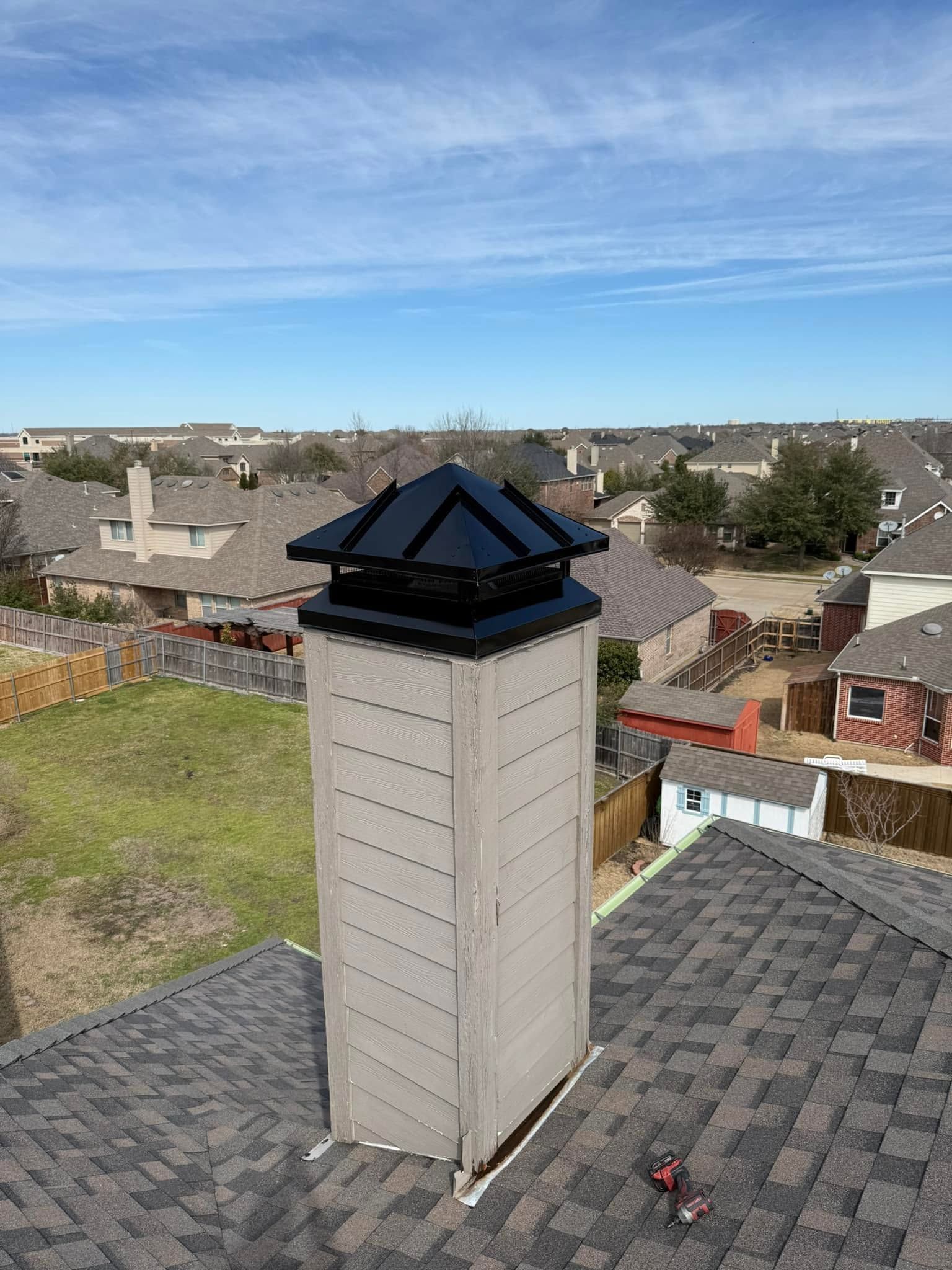 Suburban rooftop chimney with black cap under a clear blue sky, overlooking nearby houses and yards