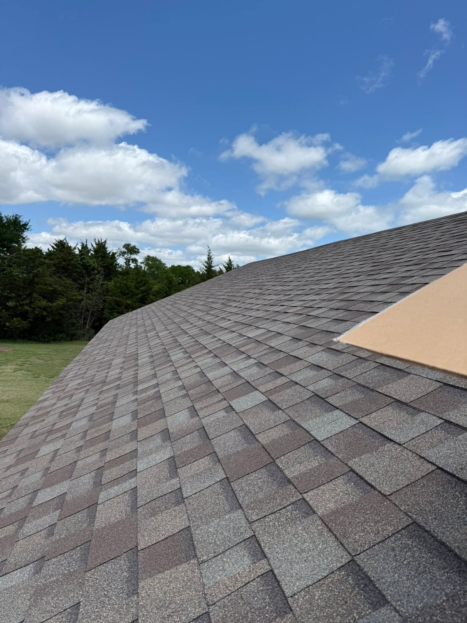 Curved gray shingle roof under a blue sky with clouds and trees beyond