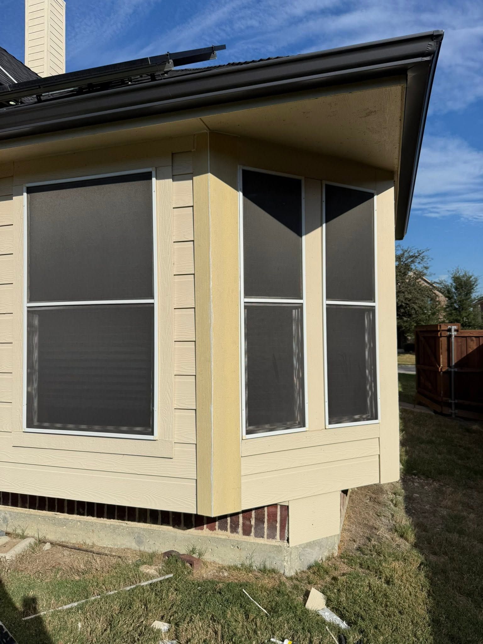 Cream-colored house corner with dark roof and large screened windows on a grassy yard.