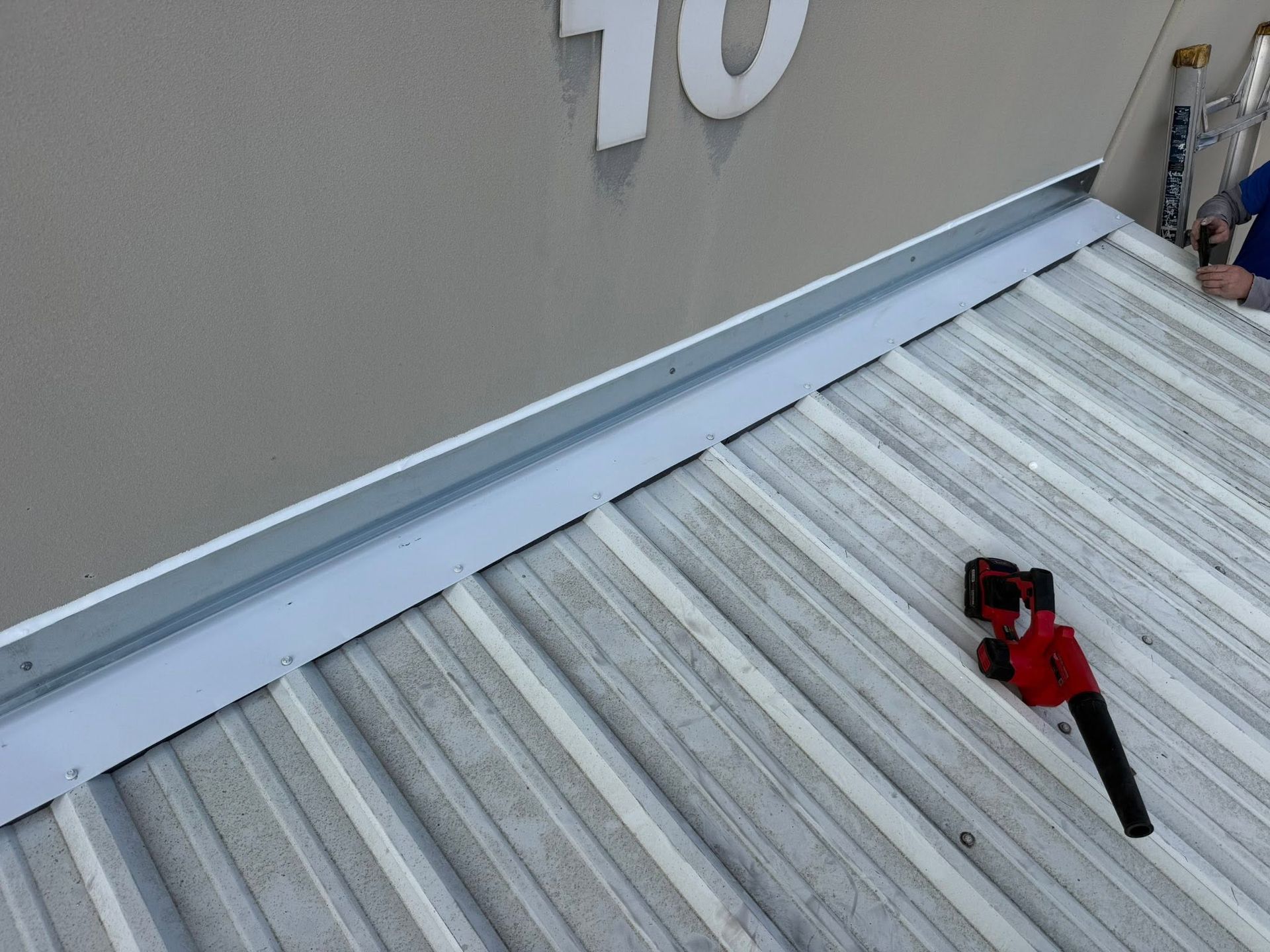 Red and black leaf blower on a corrugated metal rooftop beside a gray building wall.