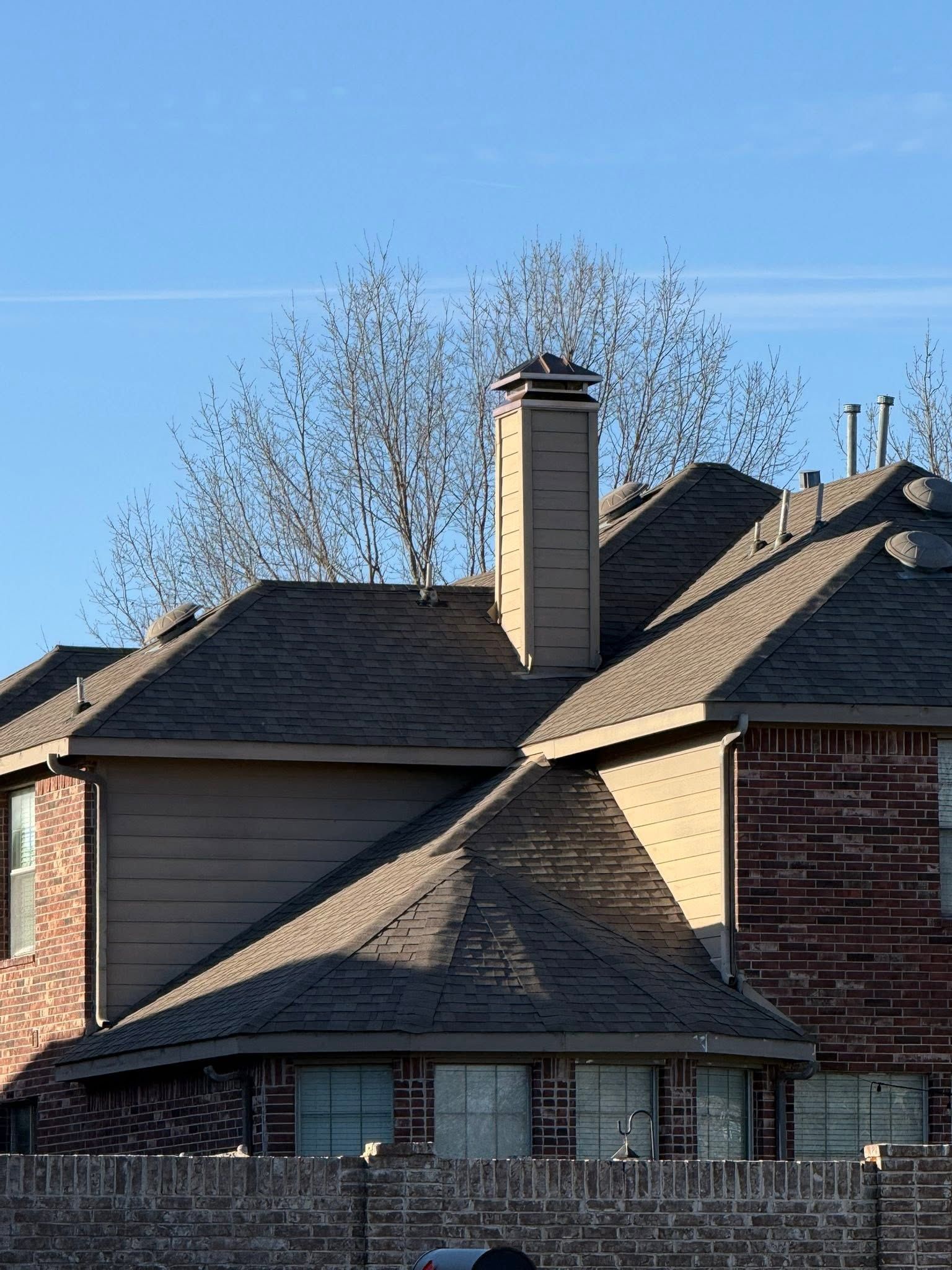 Brick house roofline with tan chimney and leafless trees against a clear blue sky