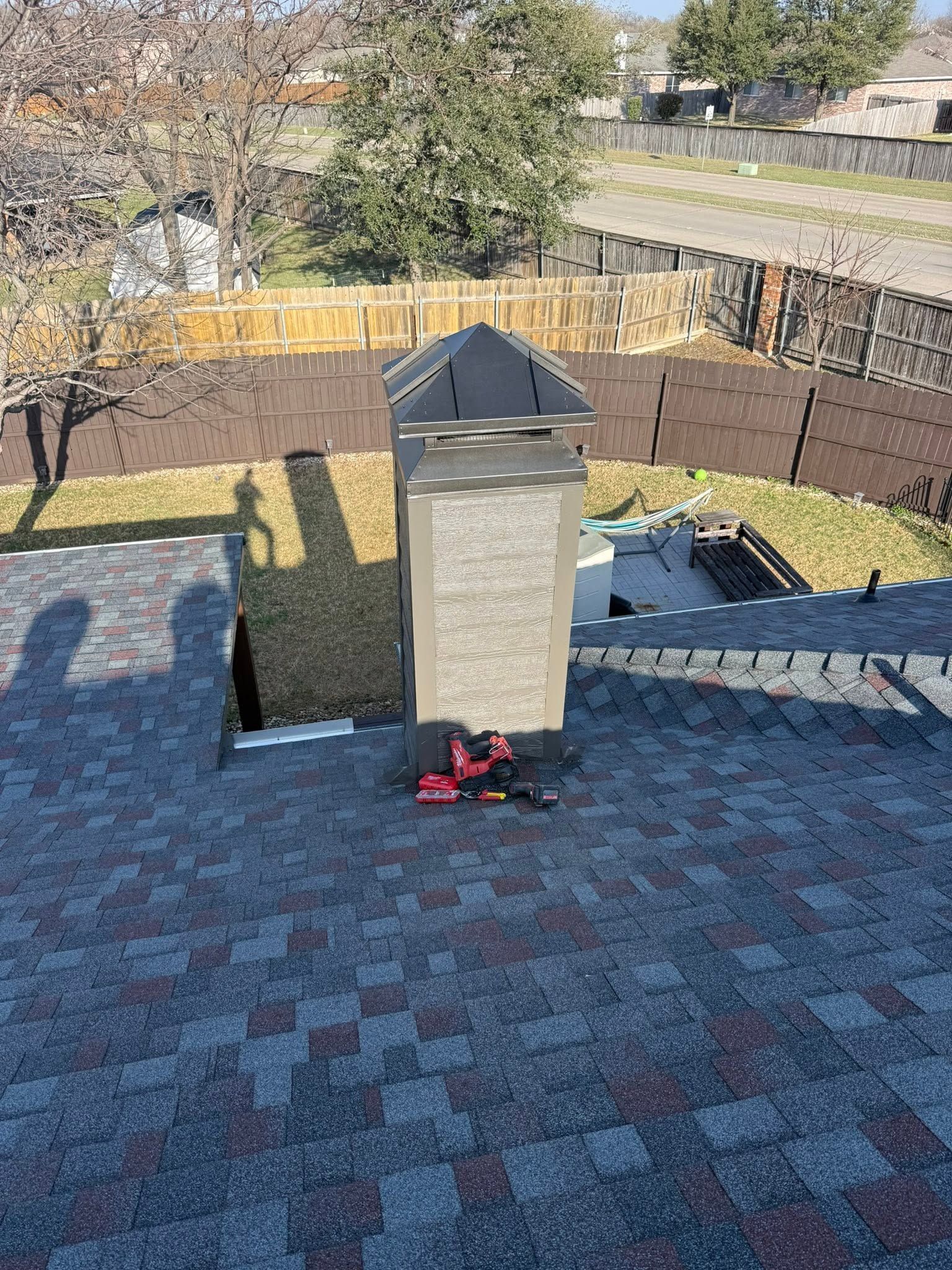 Backyard rooftop chimney with red snow blower on shingled roof, fenced yard and road beyond