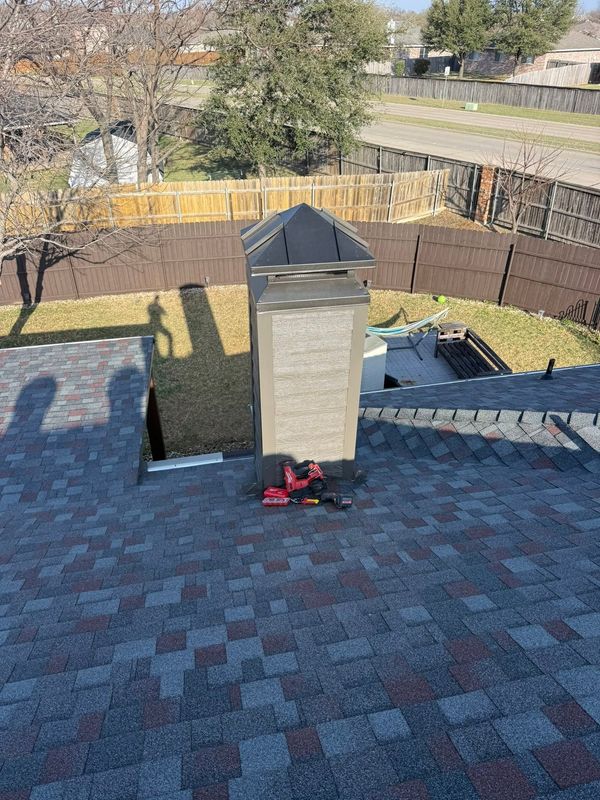 Backyard rooftop chimney with red snow blower on shingled roof, fenced yard and road beyond