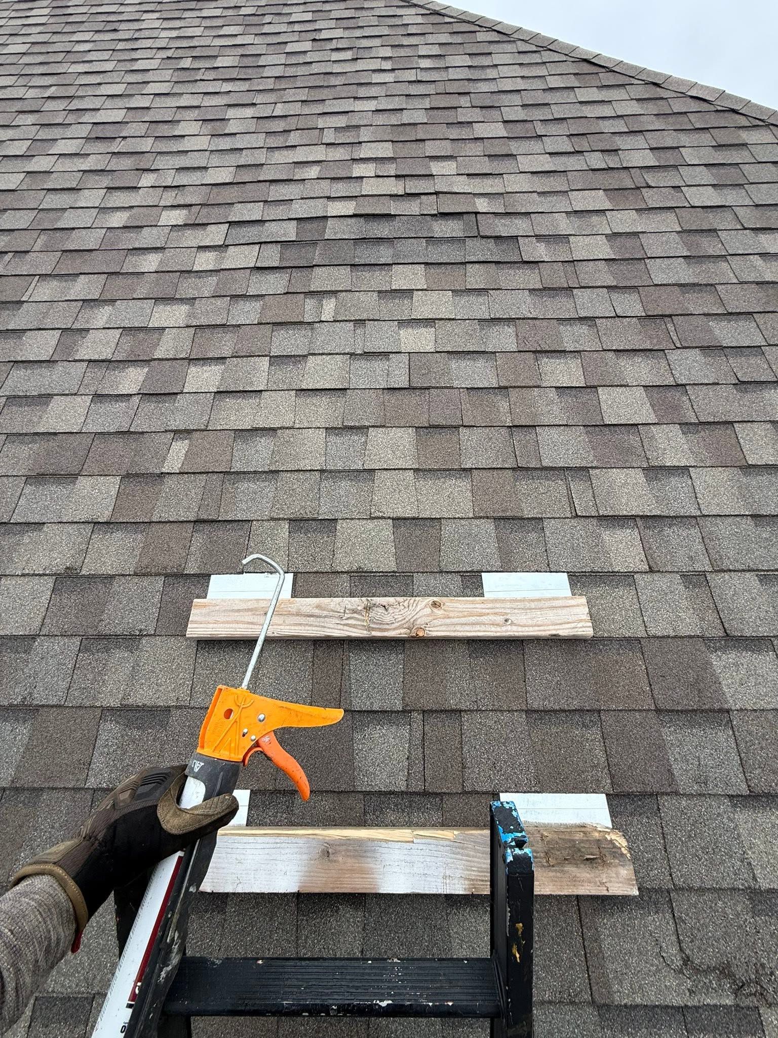 Person using an orange caulk gun on shingles near a roof ladder on a steep gray shingle roof