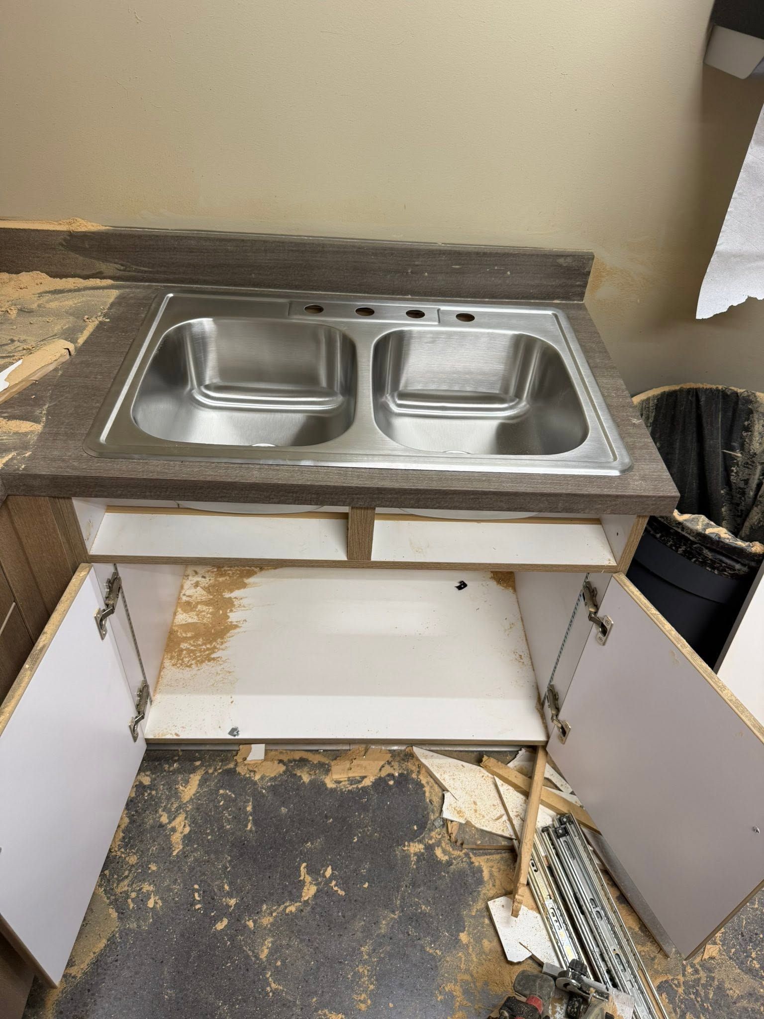 Damaged kitchen sink cabinet with double stainless-steel sink and open doors, debris scattered on the floor