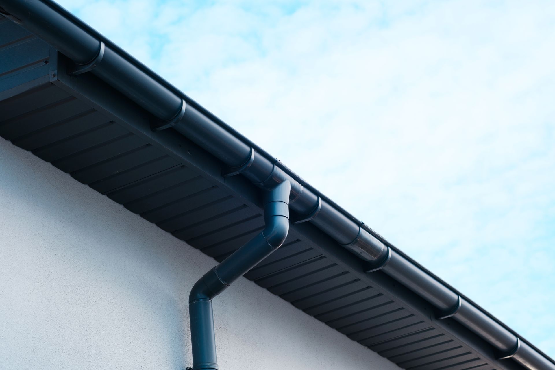 Dark blue rain gutter and downspout on a white house roof against a pale blue sky