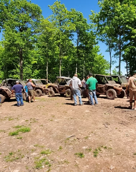 Mud-covered off-road vehicles lined up outdoors with several people standing nearby in a wooded clearing.