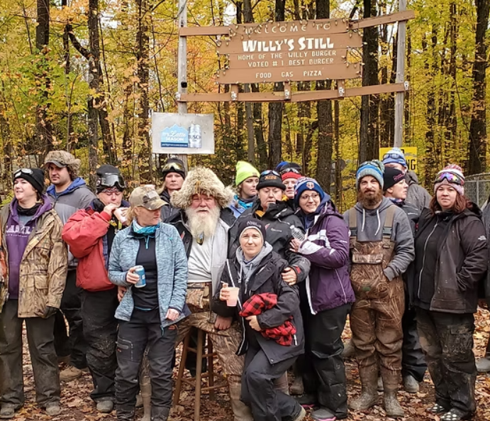 Hiking group posing at a trailhead sign in an autumn forest, holding cups and wearing jackets