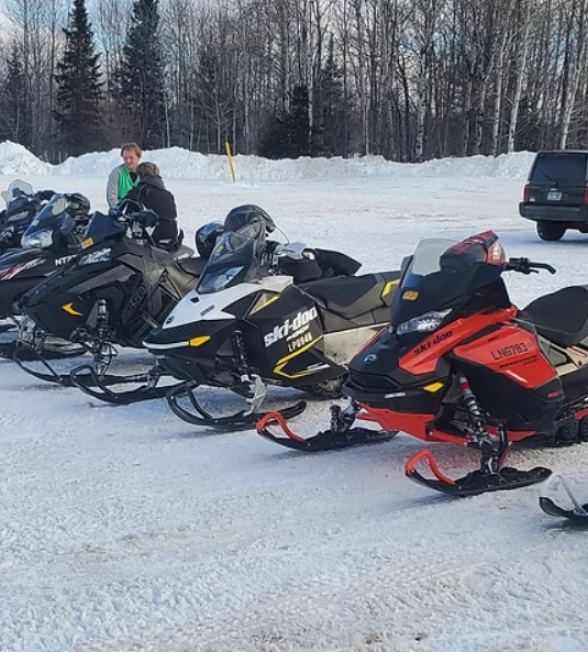 Snowmobiles lined up on snowy ground with a person standing beside them, forest in the background.