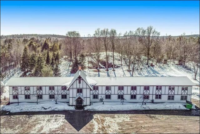 Aerial view of a long white lodge-like building with a snowy roof beside leafless trees under a clear blue sky