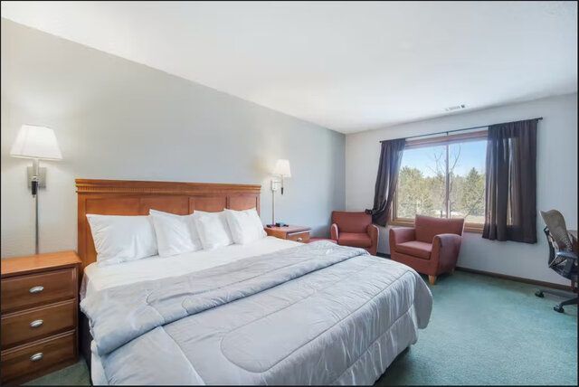 Spacious hotel room with a king bed, armchairs, lamp, and large window with curtains.