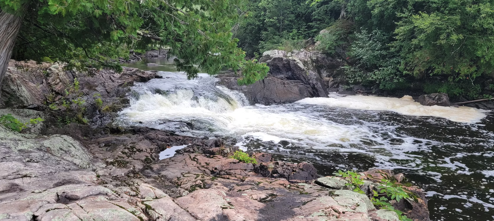 Small waterfall flowing over rocks into a shallow stream, surrounded by green foliage.