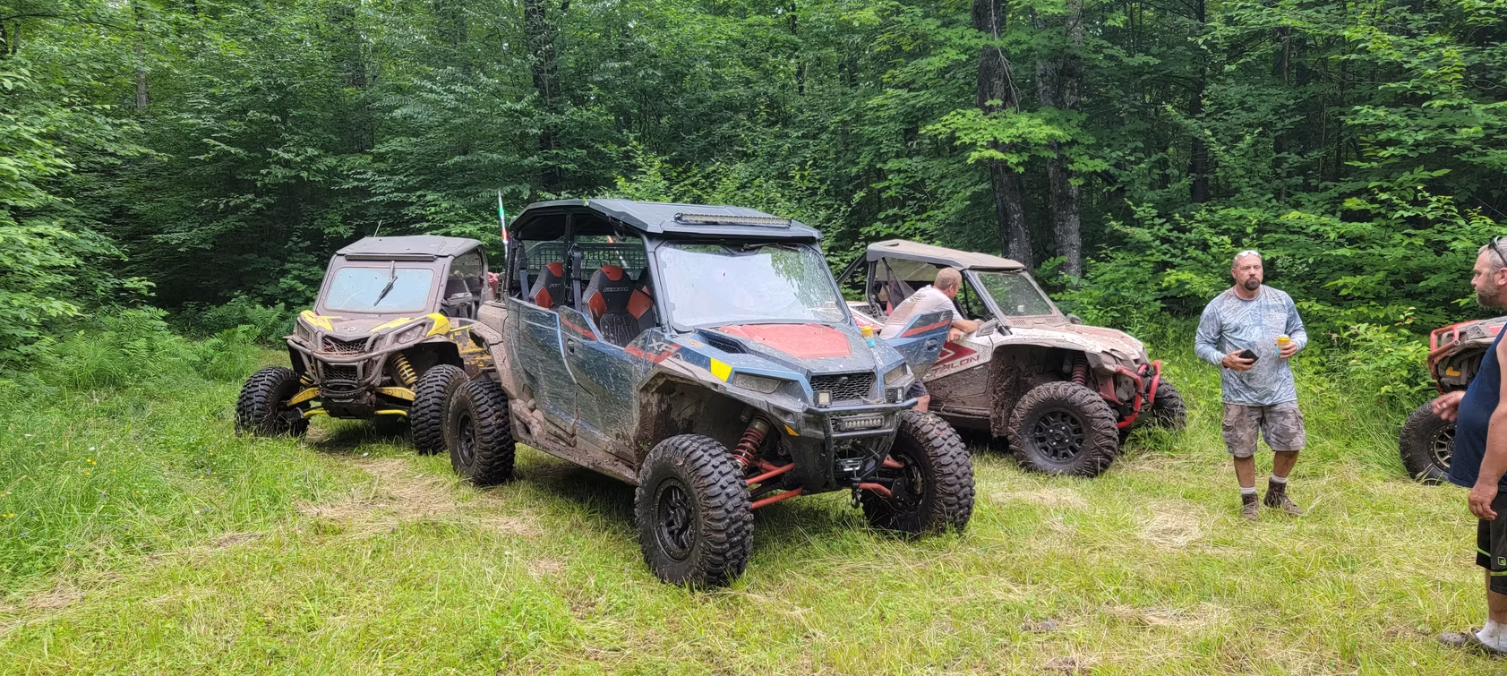 Three off-road vehicles parked in a muddy forest clearing with people standing nearby.