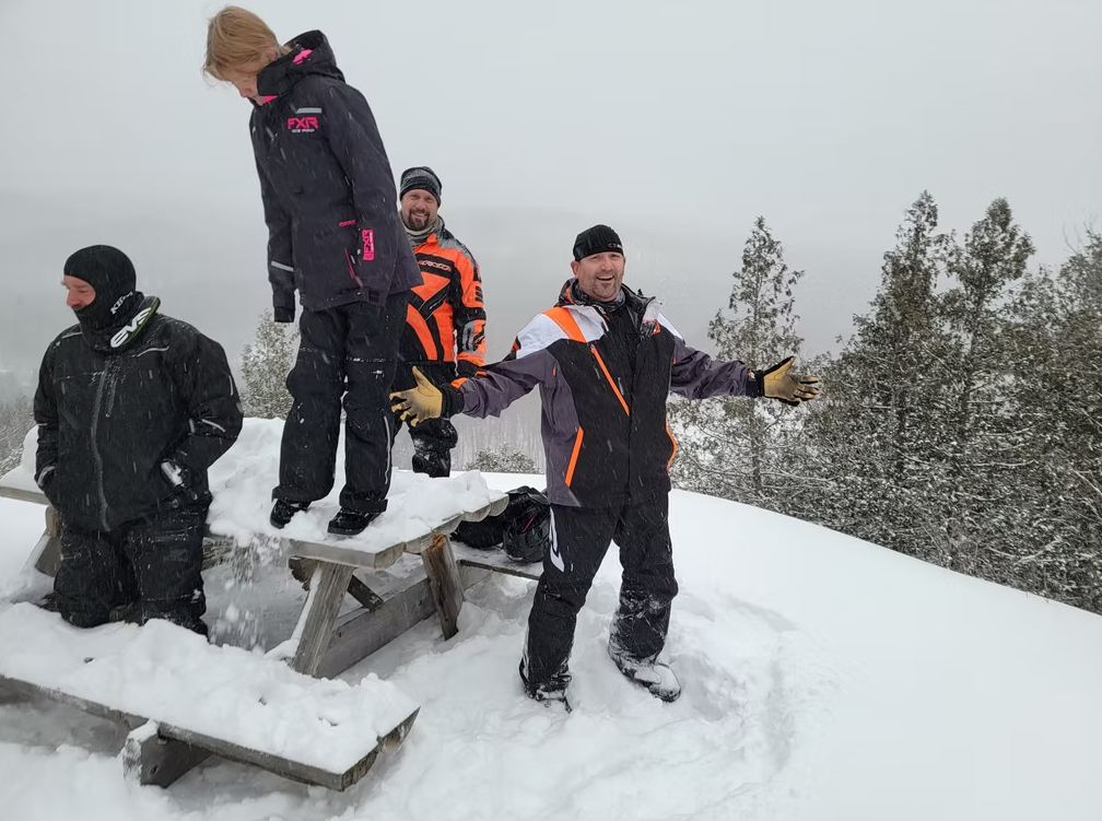 Four people in winter gear stand on a snowy hill by a bench, with trees in the background.