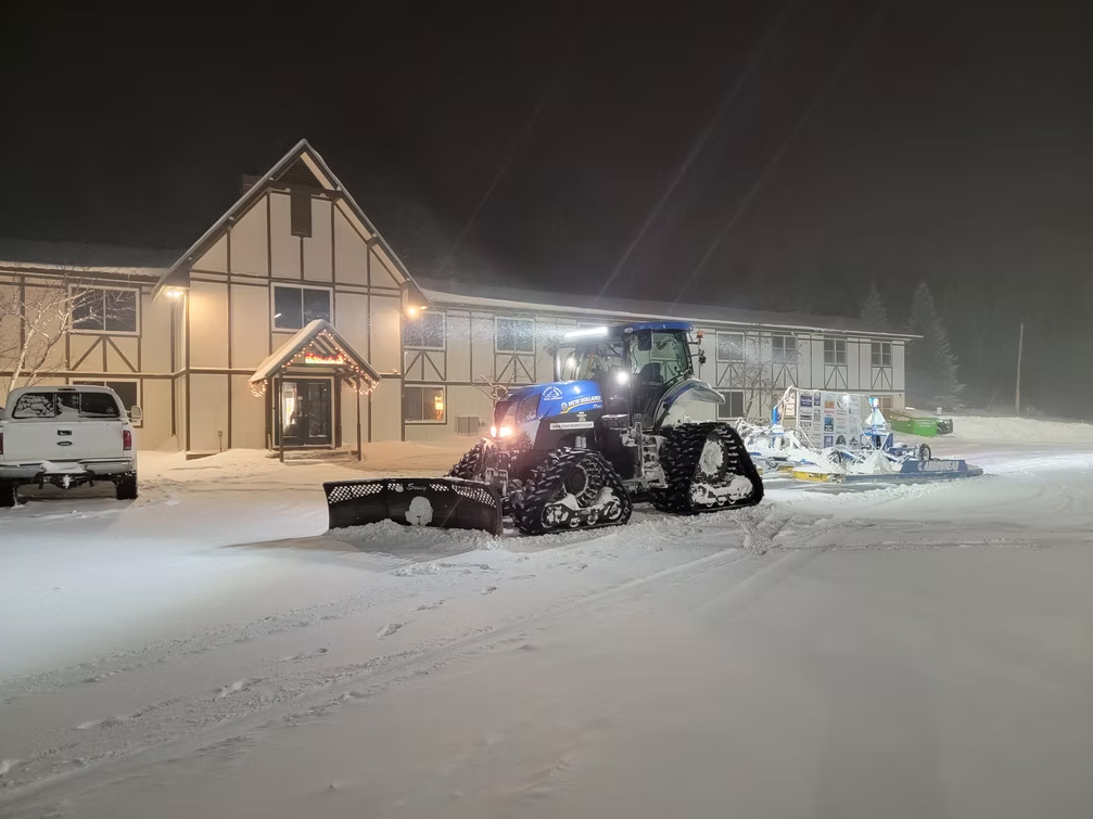 Snowplow clearing a snowy driveway at night beside a lit building
