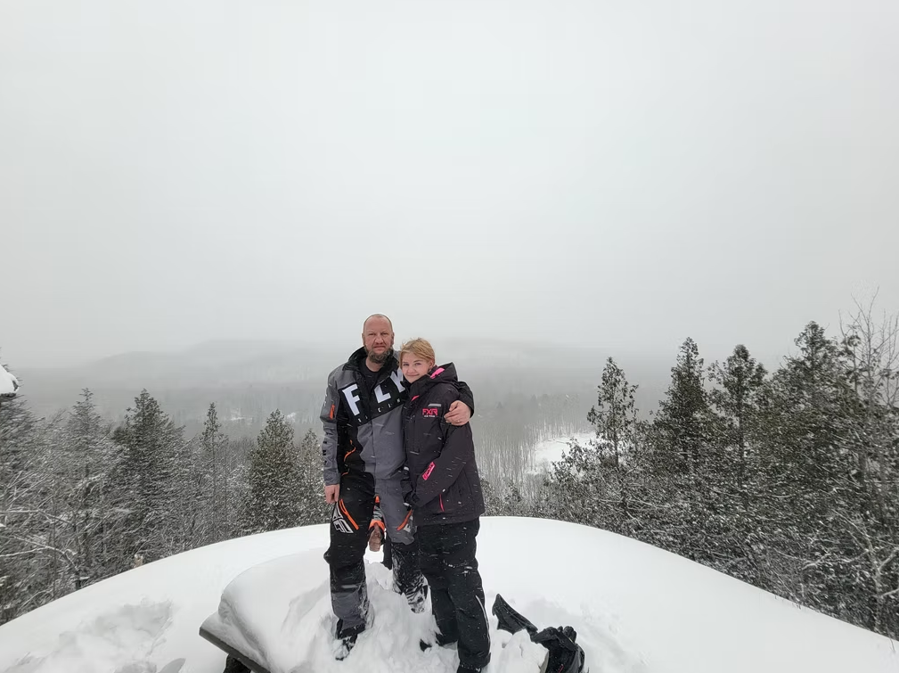 Two people standing on a snowy overlook with trees and a foggy mountain background