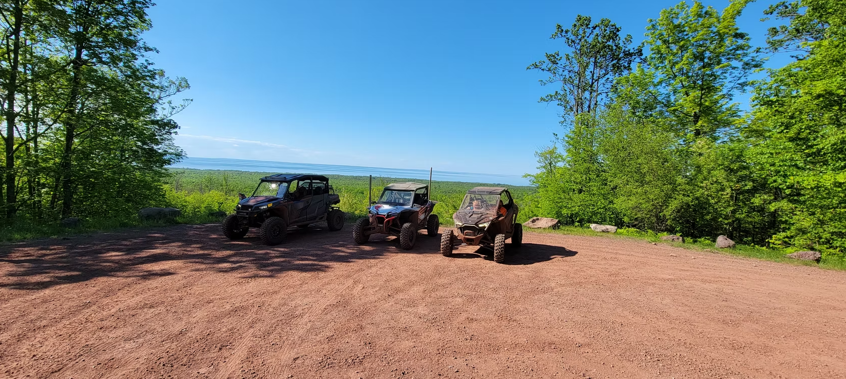 Three off-road vehicles parked on a dirt clearing with trees and a blue sky in the background