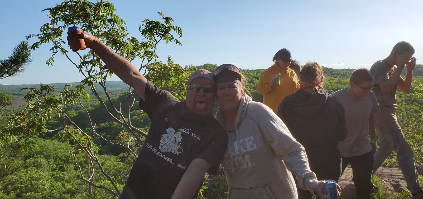 Group of people posing outdoors on a sunny hillside, smiling and gesturing with arms raised.