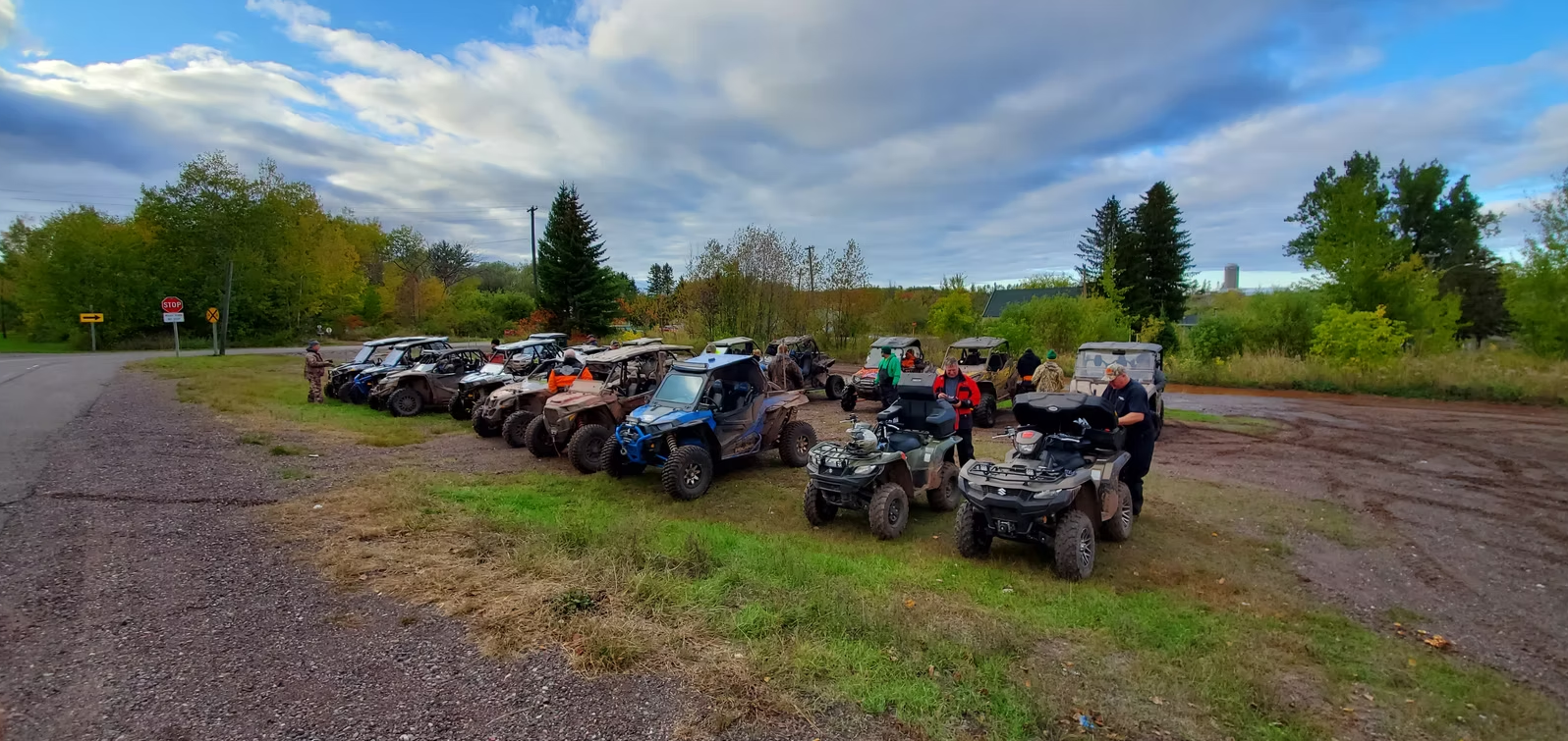 Row of parked ATVs on a gravel lot beside a wooded field under cloudy skies