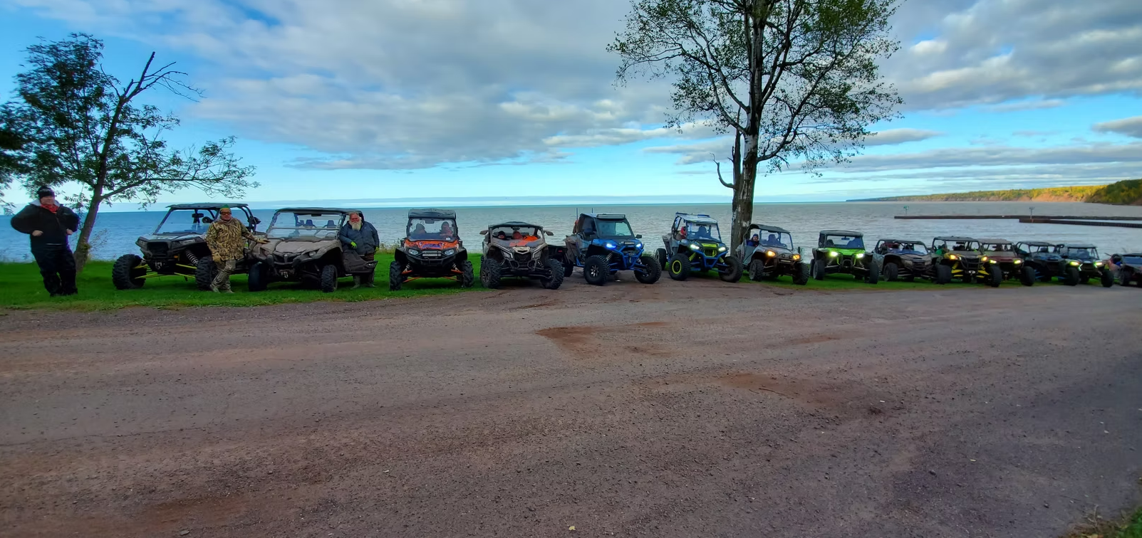 Line of off-road vehicles parked along a dirt road at dusk beneath tall trees.