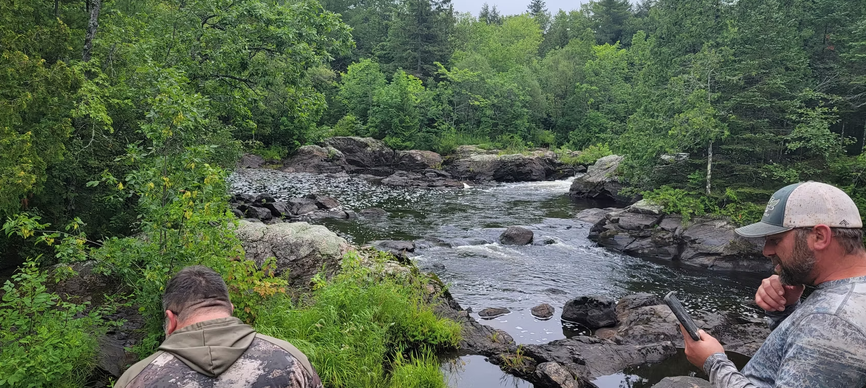 Two people sitting by a rocky river in a lush green forest, one holding a phone.